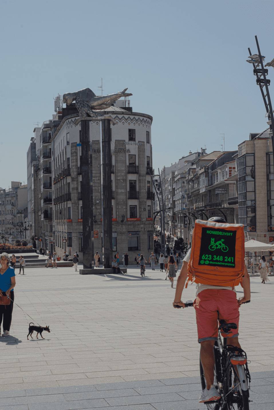 Rider de HomeDelivery con la mochila naranja pedaleando por una plaza de Vigo